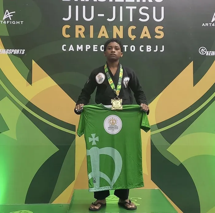 Lorran Domingos no pódio do Brasileiro de Jiu-Jitsu Crianças, de quimono preto e medalha, segurando a camisa do Instituto Imperatriz